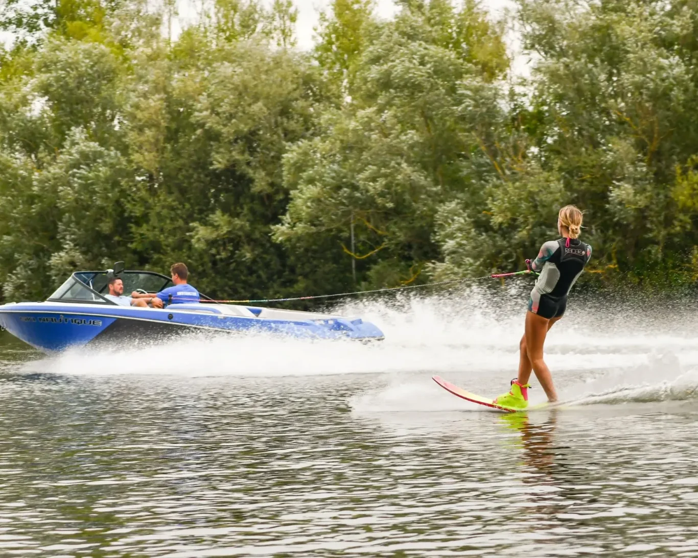 A woman water skiing behind a boat on a calm lake, surrounded by trees.