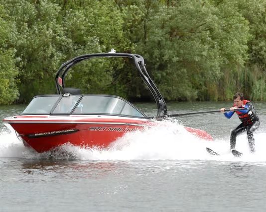A person learning to water skiing behind a red boat on a river. Green trees in the background.