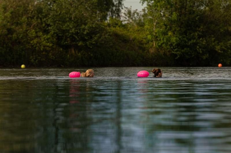 Two swimmers with pink buoys are in a calm, reflective body of water surrounded by greenery.